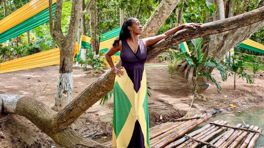 A woman in a floor-length dress with a black top and a skirt in the colors of the Jamaican flag (black, green, and gold) stands in a wooded area. She is leaning on a fallen tree trunk, with her hand on her hip and looking to the side. Yellow and green fabric ribbons are strung through the trees in the background.