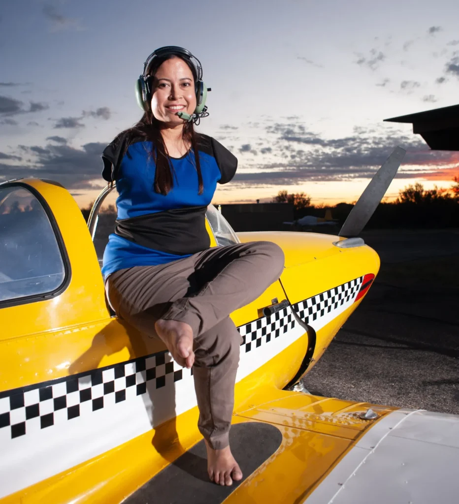 A professional portrait of Jessica Cox, a pilot born without arms, who is sitting on the wing of a yellow airplane at sunset while wearing an aviation headset.