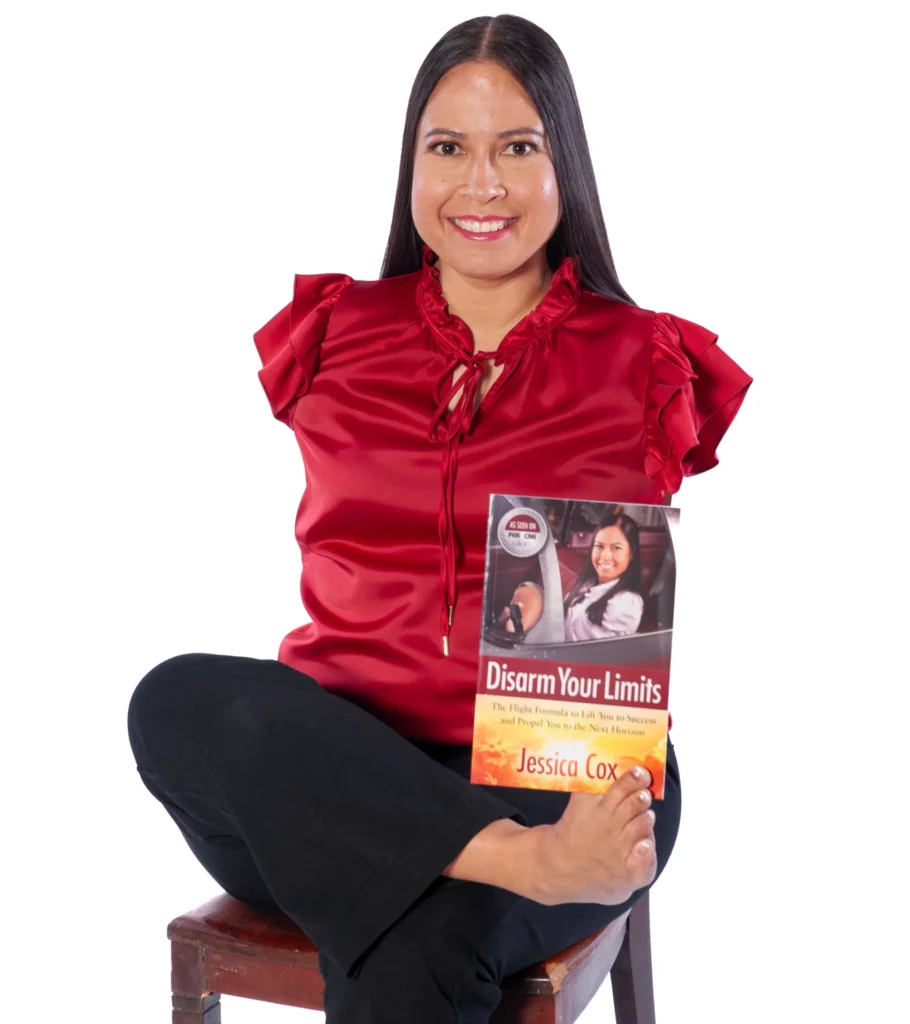 A professional portrait of Jessica Cox, a woman born without arms, who is sitting on a stool and holding her book, Disarm Your Limits, with her foot.