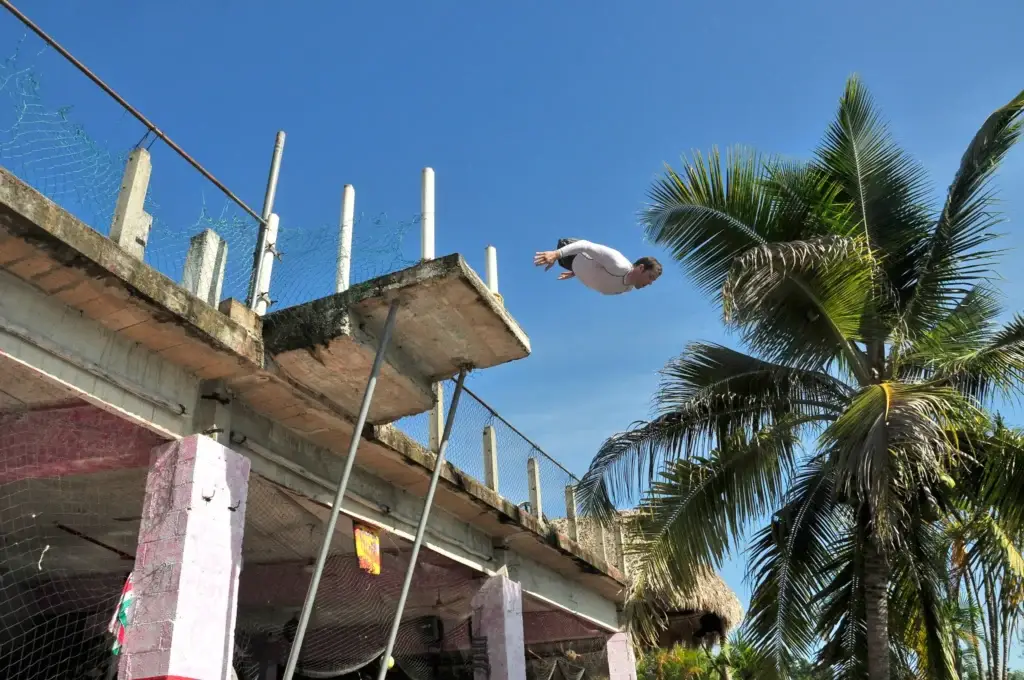 A man in a white shirt is captured in mid-air, having jumped from a concrete platform. He appears to be doing a belly flop into water below, which is not visible in the frame. The background shows a clear blue sky and a vibrant green palm tree.