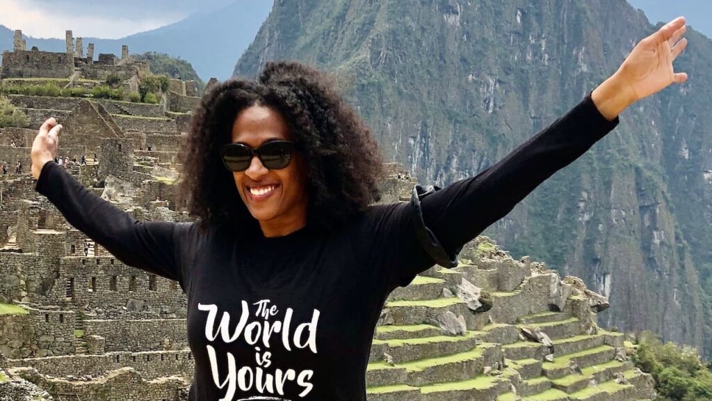 A smiling woman with curly hair and sunglasses stands in front of the ancient stone ruins of Machu Picchu, with her arms outstretched. She is wearing a black t-shirt that says "The World is Yours" in white script.