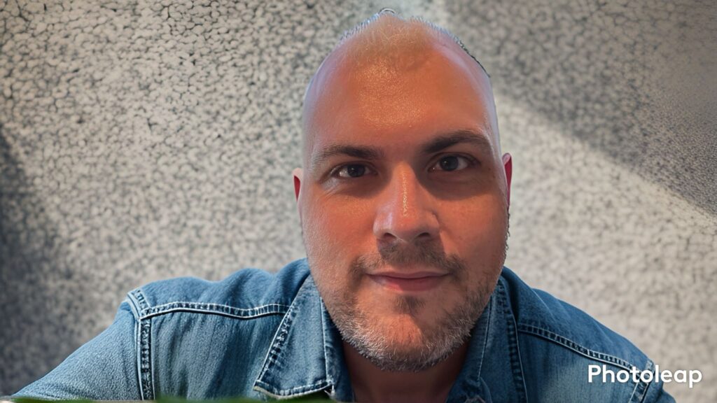 A close-up selfie of a man with a shaved head and a light beard, wearing a denim shirt, looking directly at the camera. The background is a blurred, gray, textured surface.