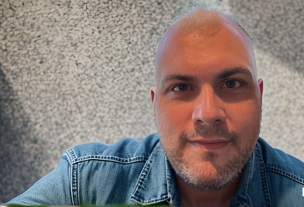 A close-up selfie of a man with a shaved head and a light beard, wearing a denim shirt, looking directly at the camera. The background is a blurred, gray, textured surface.