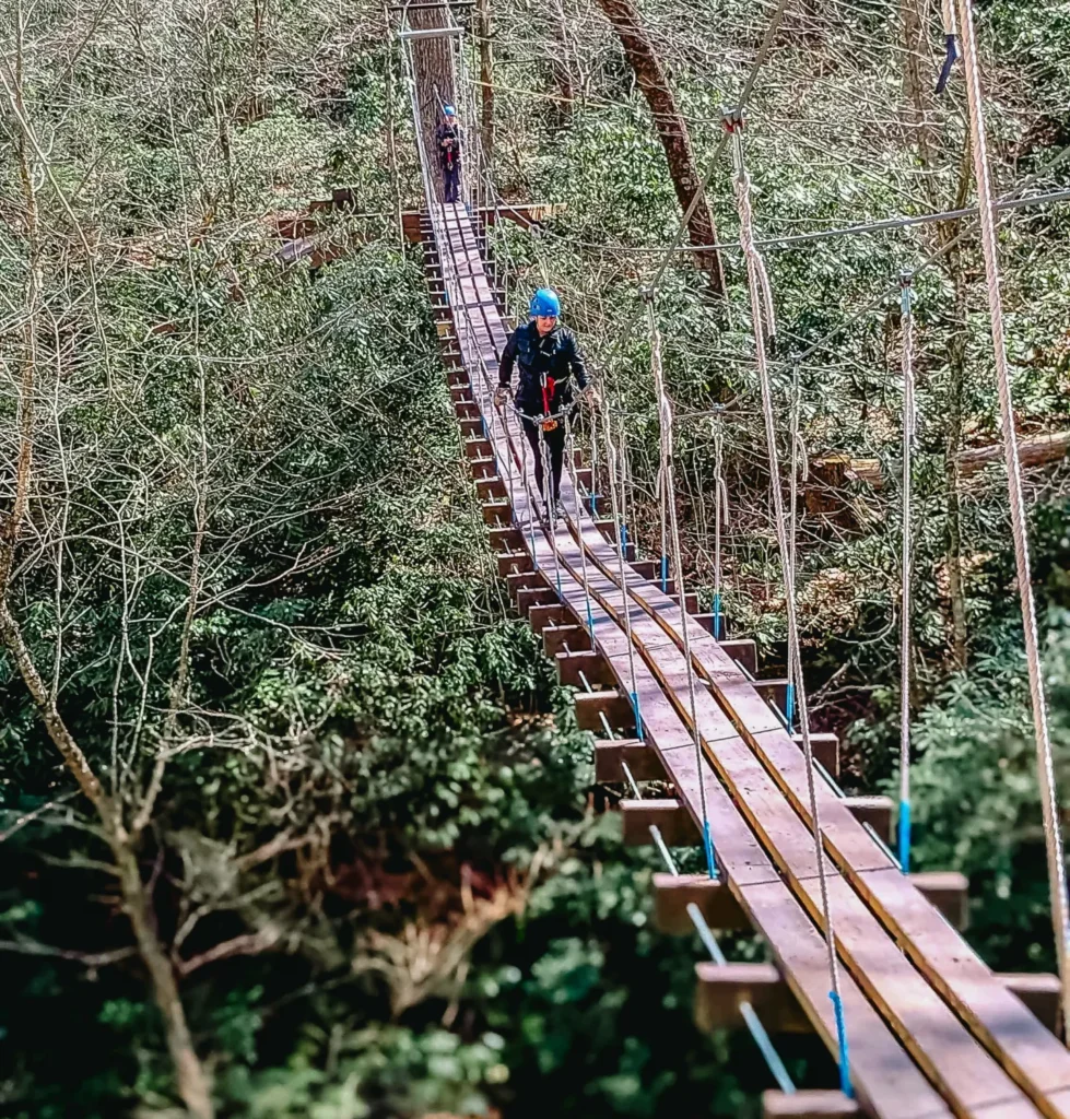 A person wearing a helmet walks across a wooden and rope suspension bridge high above a rocky riverbed in a forest.