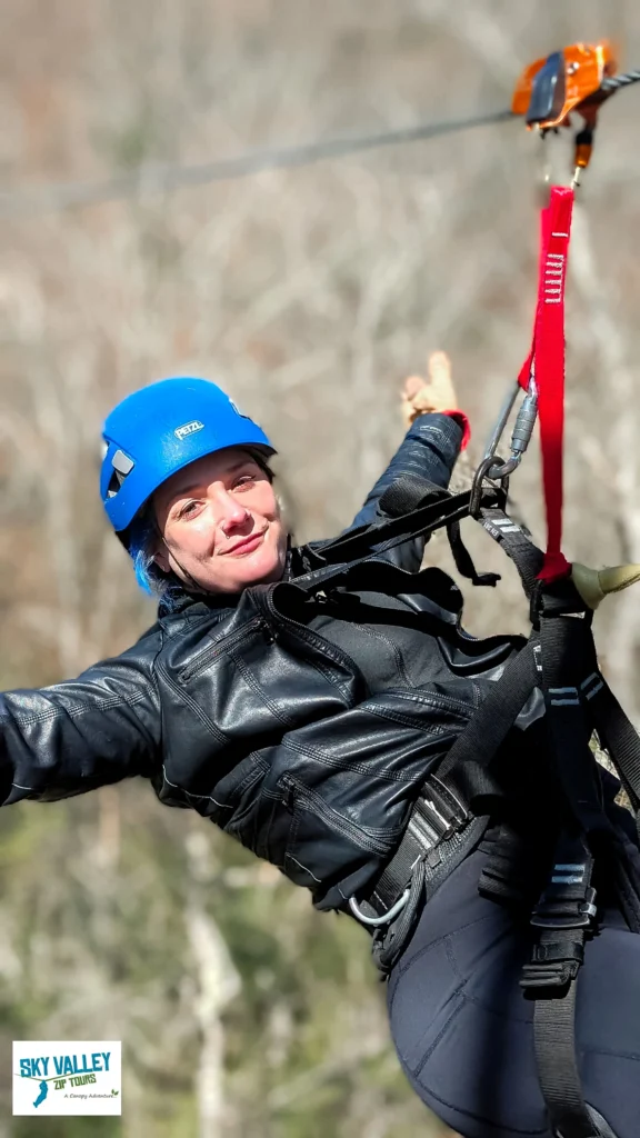 A woman wearing a blue helmet and a black jacket smiles and gives a thumbs-up while riding a zipline.
