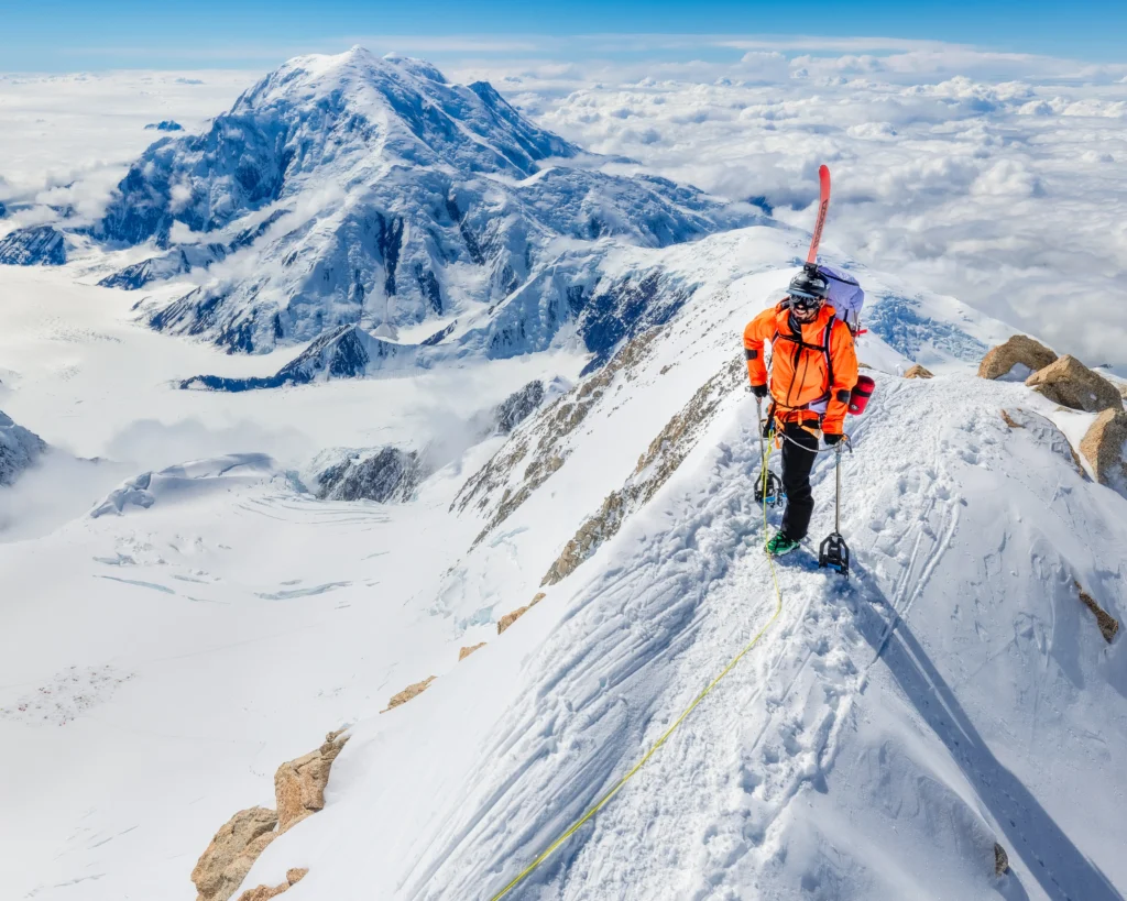 A man in a bright orange jacket and helmet is hiking on a narrow, snow-covered ridge. He has skis attached to his backpack and is using an ice axe. The background shows a massive, snow-covered mountain peak under a clear blue sky, with clouds filling the valleys below.