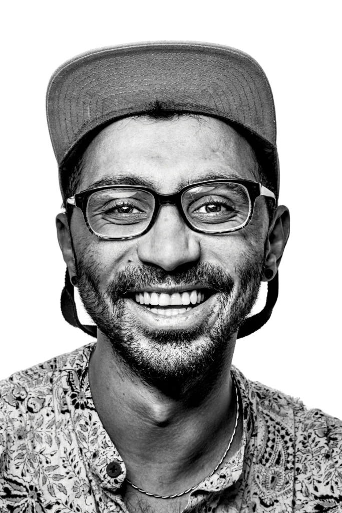 A close-up, black-and-white portrait of a man with a wide smile, wearing glasses and a backward baseball cap
