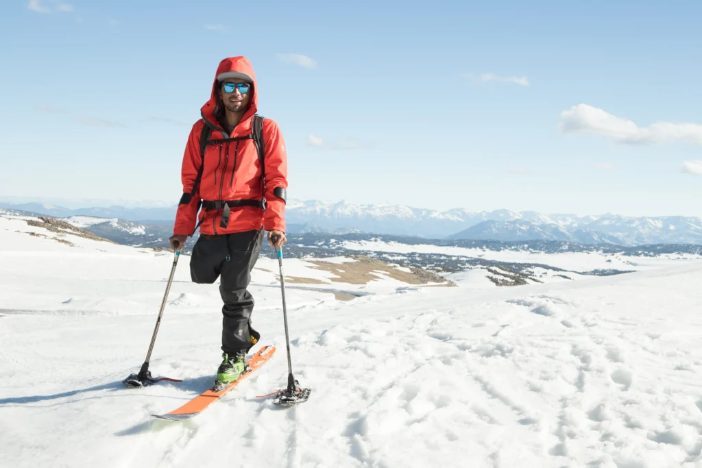 A man in a red jacket, dark pants, and a baseball cap standing on skis in a vast, snow-covered landscape. He is holding ski poles and wearing sunglasses. The background shows rolling hills and distant mountains under a blue sky with scattered clouds.