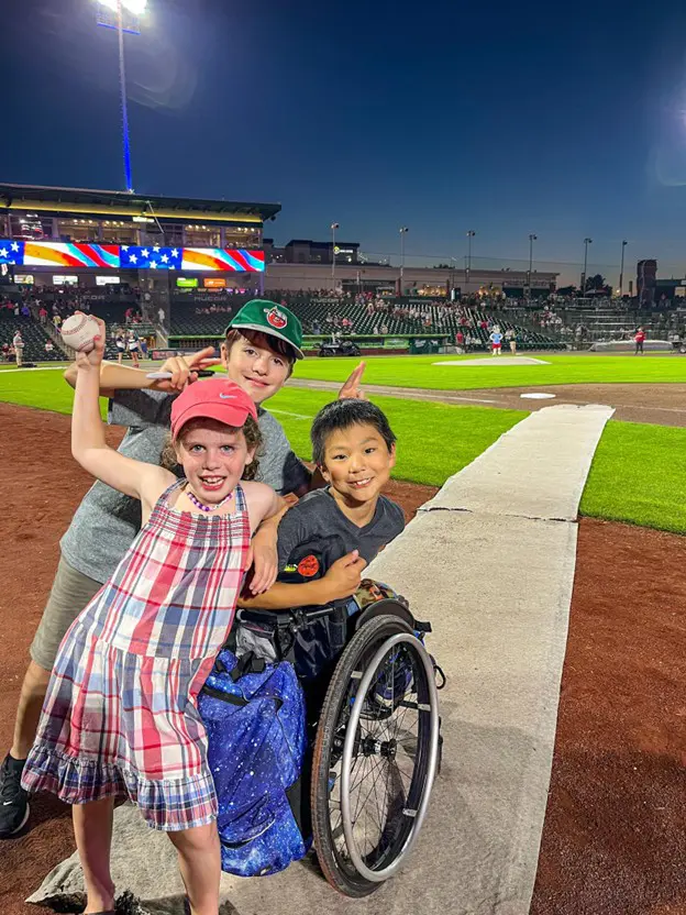 Three children are on the grass beside a baseball field at dusk. The girl in the foreground holds a baseball and is looking at the camera. A boy in a wheelchair and another boy with a baseball cap are beside her.