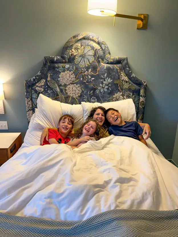 A woman and three children are lying in a bed with white covers and a large headboard with a floral design. All of them are smiling and looking at the camera.
