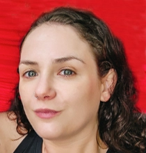 A headshot of a woman with brown hair and green eyes, smiling slightly. She is wearing a black top and a red background is visible behind her.