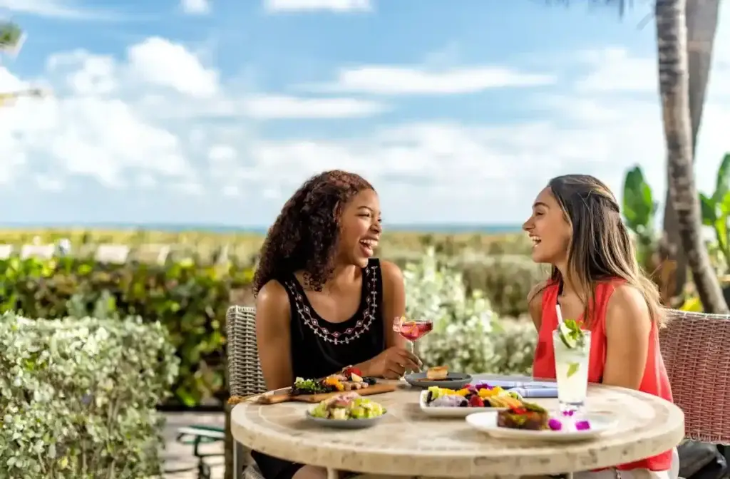 Two women are laughing and talking while seated at an outdoor restaurant table with the ocean in the background. They are sharing a meal and drinks.