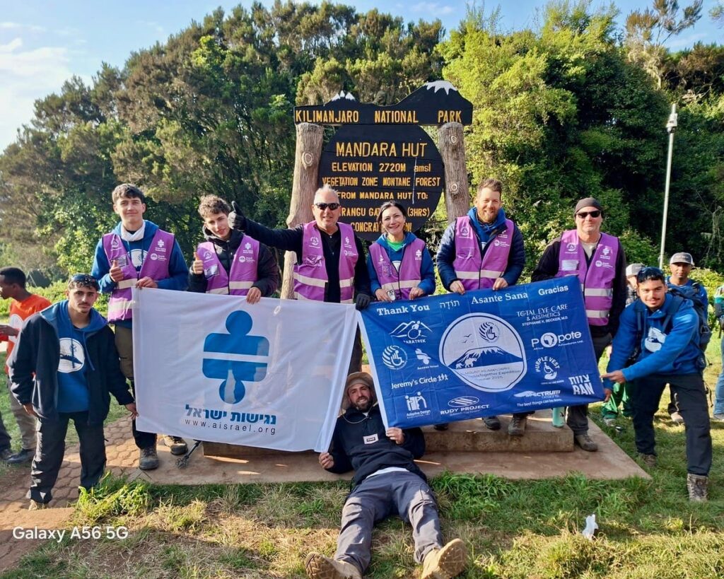 A group of hikers, some in purple vests, poses with flags in front of a wooden sign for Mandara Hut in Kilimanjaro National Park. One person is lying on the ground in front, holding a flag.