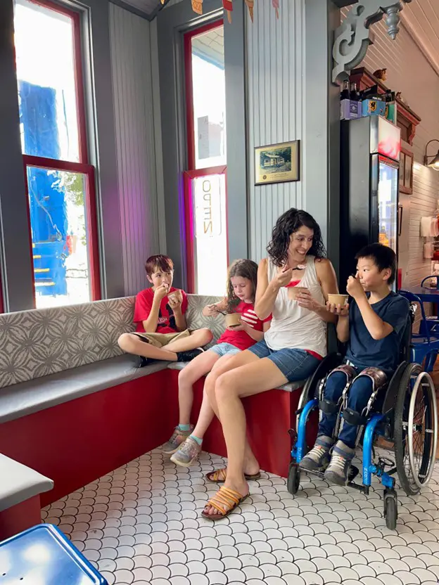 A woman and three children are sitting in an ice cream shop eating ice cream. The woman and a girl are on a red and gray bench, and two boys are in chairs across from them. One of the boys is in a wheelchair.