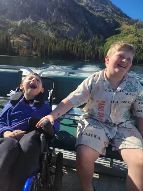 Two boys are on a boat with a lake and mountains in the background. The boy on the left is in a wheelchair and is laughing with his mouth wide open. The boy on the right is also laughing and leaning toward the boy in the wheelchair.