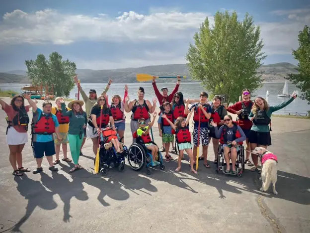 A large group of people, including several in wheelchairs, poses for a group photo near a lake. Many of them are wearing life jackets and some are holding paddles. A golden retriever stands on the right.