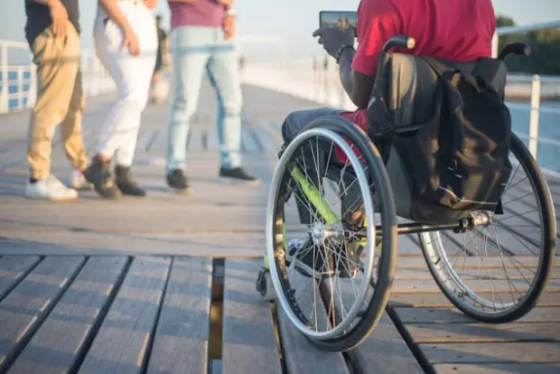 A person in a wheelchair, seen from behind, is on a wooden pier holding a tablet. In the background, three people are walking.