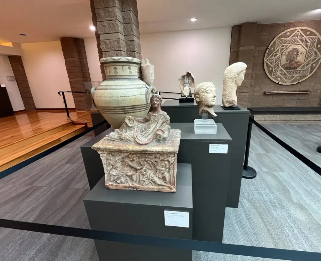 Inside a museum, a variety of ancient-style statues and a large clay vase are displayed on elevated gray platforms. The background features light-colored walls and a wooden floor. The pieces appear to be from different historical periods.