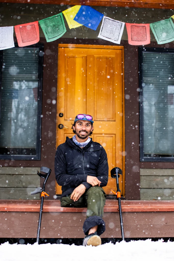 A man with a prosthetic leg is sitting on a porch. He is wearing a dark jacket, and there are two crutches on either side of him. The porch has a wooden door and prayer flags hanging above it. It is snowing lightly.