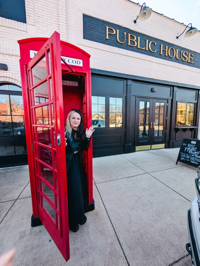 A woman with black and blonde hair waves from inside a classic red telephone booth. She is wearing a black outfit. The telephone booth is on a sidewalk in front of a brick building with a sign that reads "PUBLIC HOUSE."
