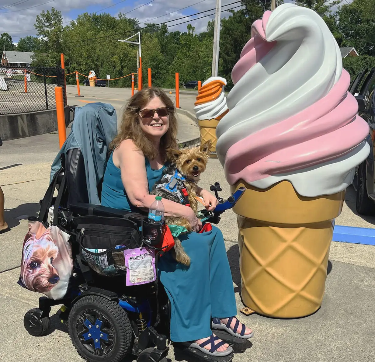 A smiling woman with sunglasses sits in a motorized wheelchair next to a large, colorful statue of a pink-and-white soft-serve ice cream cone. She is holding a small brown and black Yorkshire terrier. In the background, there is another ice cream cone statue and a drive-thru building.