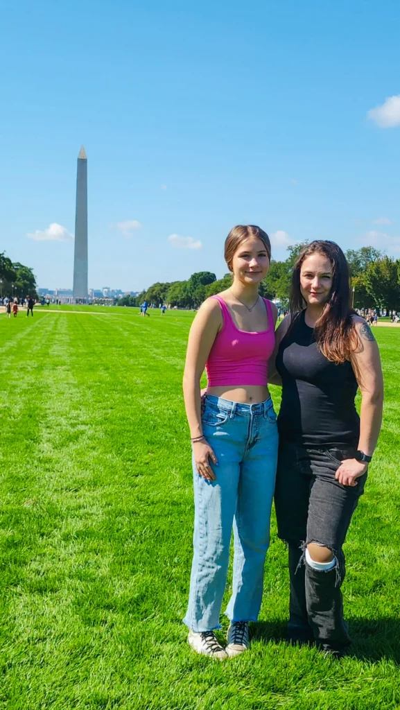 Two women stand together in a grassy field, posing for a photo with the Washington Monument in the background.