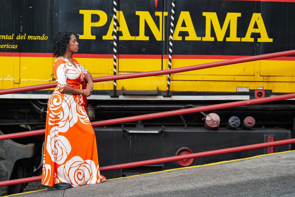 A woman in a vibrant orange and white floral dress stands on a train platform, facing left and looking into the distance. Behind her is a black and yellow train engine with the word "PANAMA" written in large yellow letters.