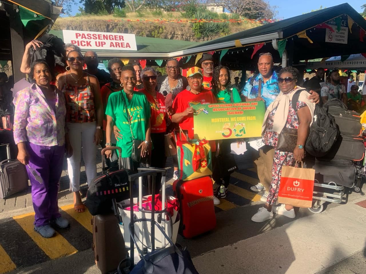 A group of people stands together outside an airport terminal with their luggage. They are holding a large sign that says, "DISCOVER YOUR ROOTS GRENADA/CARRIACOU AND PETITE MARTINIQUE ASSOCIATION/QUEBEC (DYRGAQ) MONTREAL WE COME FROM."