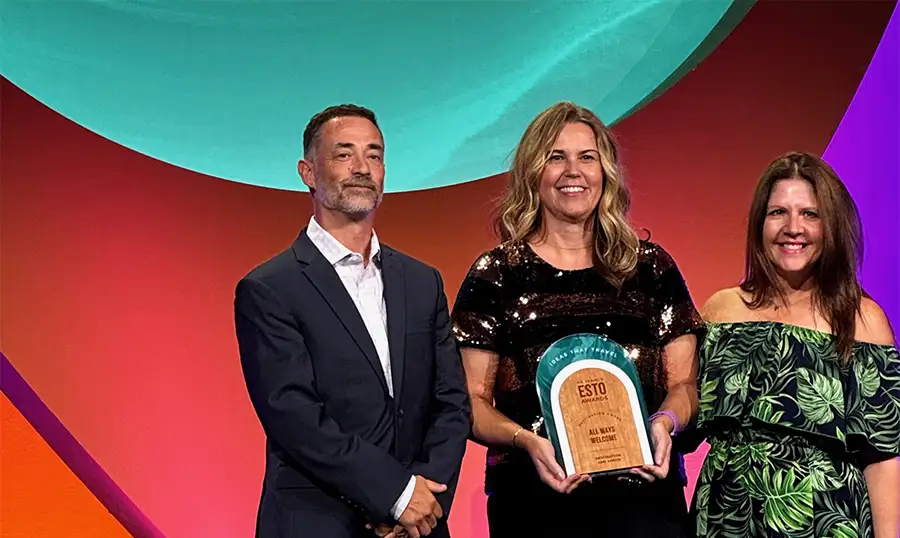 A man and two women stand on a stage in front of a large screen displaying "U.S. TRAVEL'S ESTO AWARDS." The woman in the center is smiling and holding a plaque that reads "All Ways Accessible."