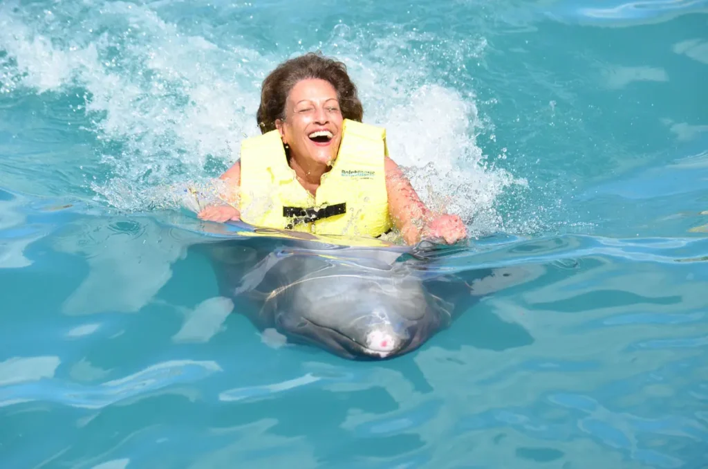 A smiling woman in a yellow life vest is holding a dolphin in the water.