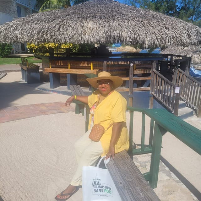 A woman in a straw sun hat and a yellow button-up shirt sits on a wooden railing on a sandy beach. A thatched-roof hut is behind her. She's holding a white tote bag with a French phrase printed on it.