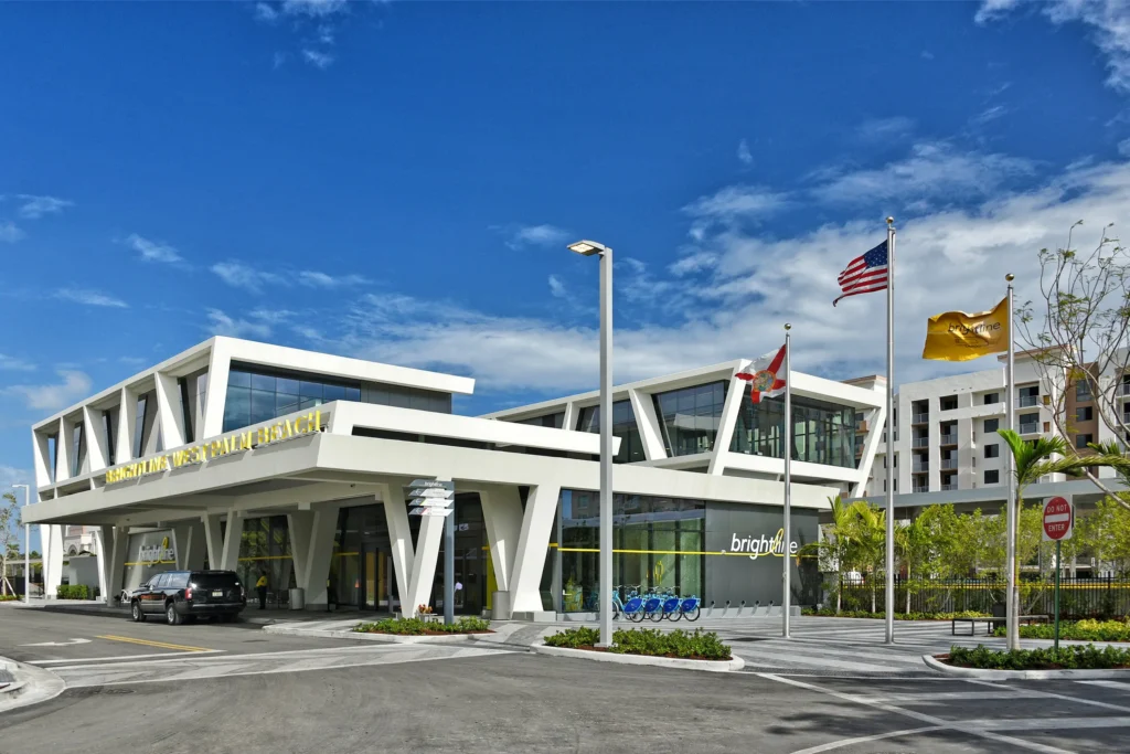 A modern, white geometric train station building under a bright blue sky. The structure features sharp angles and a large overhanging roof. Three flags—the American flag, the Florida state flag, and a yellow Brightline flag—fly on tall poles in front of a paved plaza.