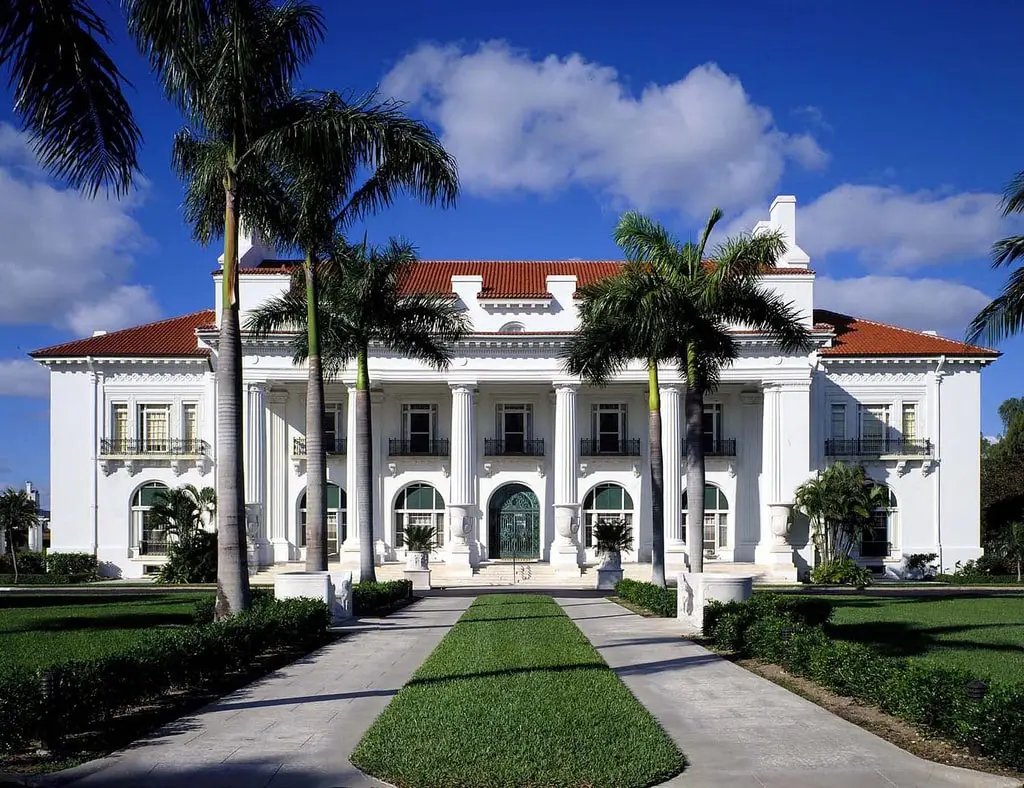 A grand, white Gilded Age mansion with large Neoclassical columns and a red-tiled roof. A symmetrical concrete path lined with green grass and manicured hedges leads to the main entrance, flanked by tall palm trees against a clear blue sky.