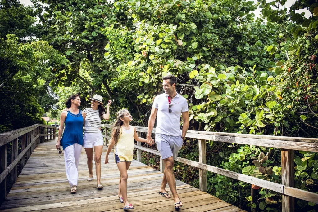 A family of four—two women, a man, and a young girl—smiling and walking together across a wooden boardwalk. They are surrounded by lush, thick green tropical foliage under bright sunlight.
