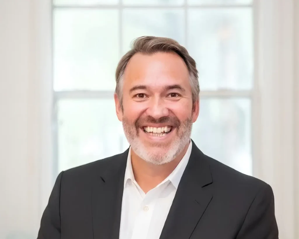 A professional close-up headshot of a man with short brown and gray hair and a neatly trimmed beard. He is wearing a dark blazer over a crisp white button-down shirt and has a warm, friendly smile. The background is a soft-focus view of a white-framed window.
