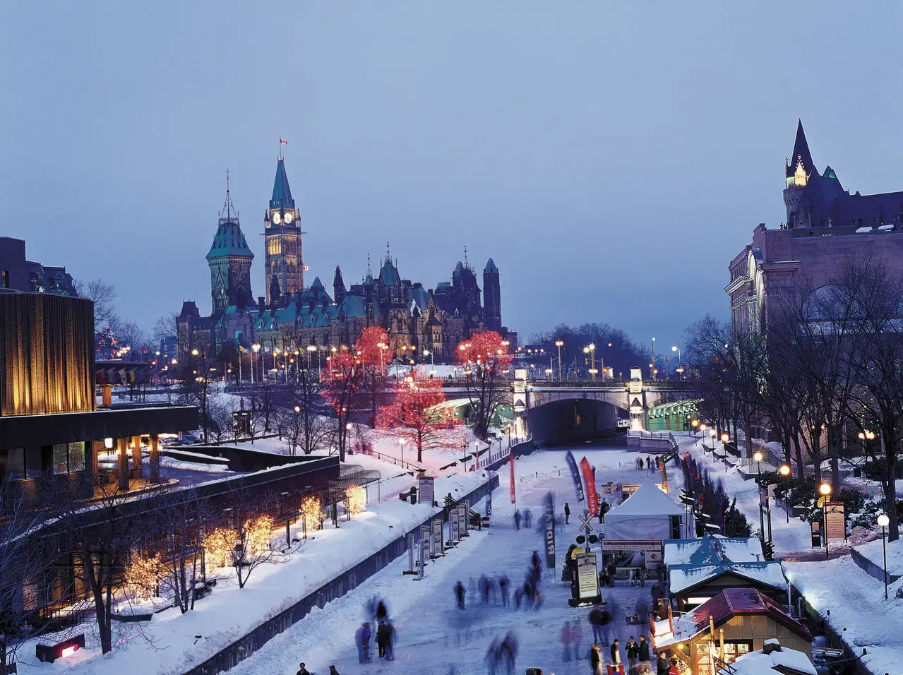 A high-angle evening view of the Rideau Canal Skateway in Ottawa during winter. The frozen canal is filled with the blurred motion of ice skaters, flanked by snowy banks illuminated by warm golden streetlights and red-lit trees. In the background, the Gothic-style architecture of Parliament Hill and the Peace Tower stands out against a deep blue twilight sky.