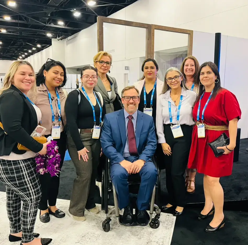 A man in a blue suit and burgundy tie, using a manual wheelchair, is positioned in the center of a group of eight professionally dressed women. They are all smiling at a trade show or conference event, identifiable by the blue lanyards and ID badges they are wearing.