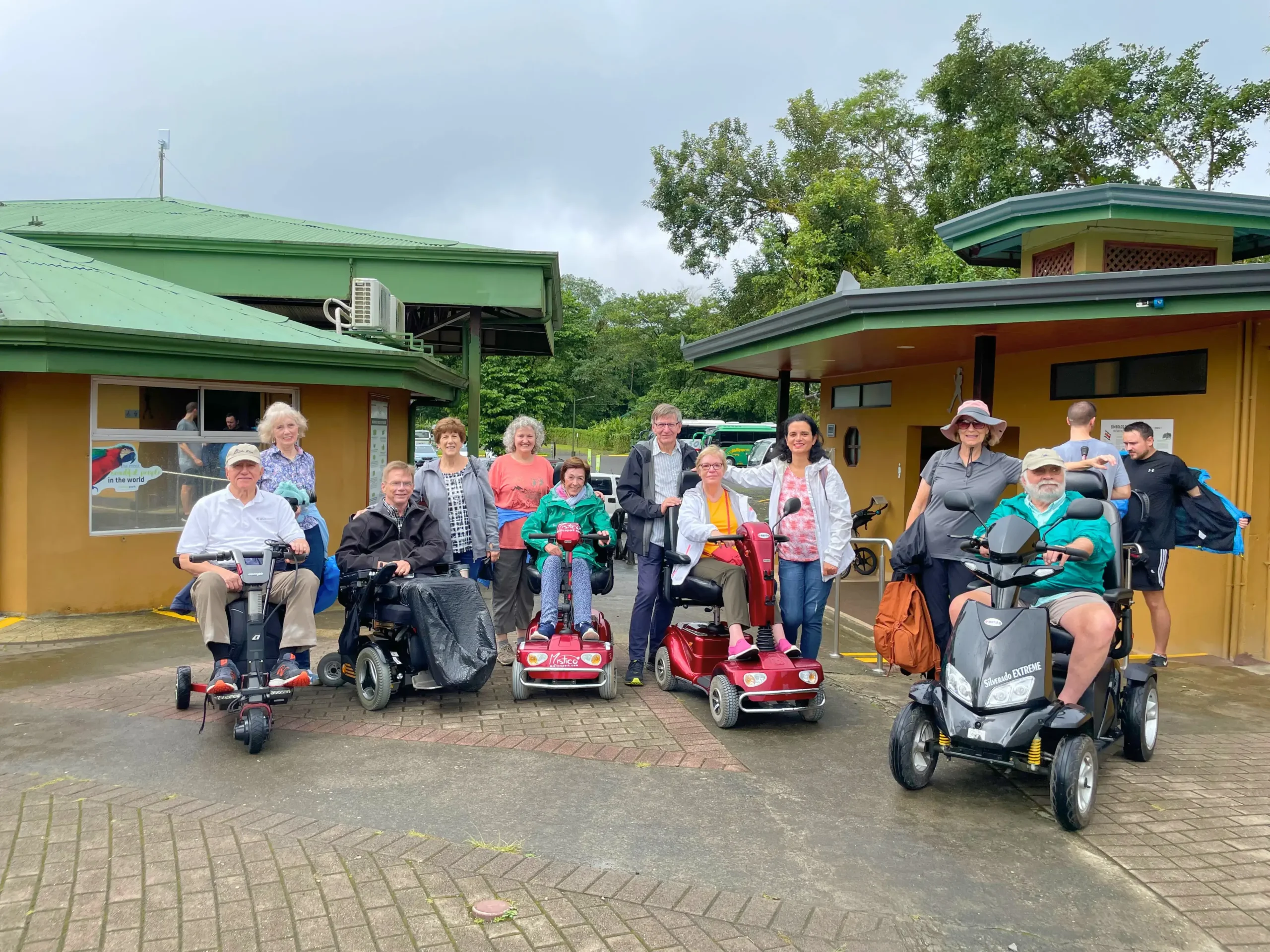 A group of ten travelers, including several individuals on red and black mobility scooters, gathered in front of tropical, green-roofed buildings in Costa Rica. The setting is lush and green under an overcast sky, with the group dressed in casual travel attire and light jackets.