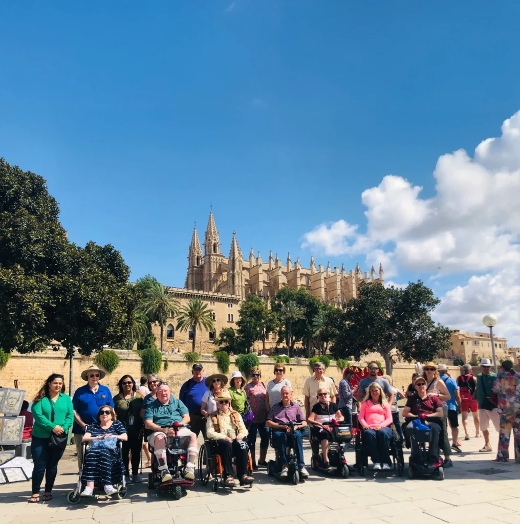 A wide group shot of approximately twenty travelers, several of whom are using wheelchairs and mobility scooters, posing on a sunny stone plaza in Mallorca. In the background, the majestic Gothic architecture of the Santa Maria of Palma Cathedral rises against a bright blue sky with scattered white clouds.