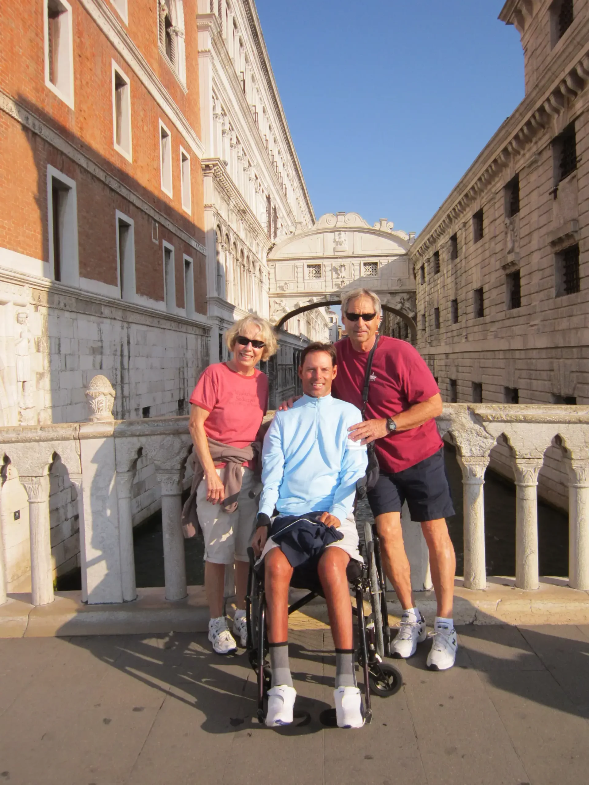A man in a light blue long-sleeved shirt sitting in a manual wheelchair, flanked by a woman in a pink shirt and a man in a maroon shirt. They are posing on a stone bridge in Venice, Italy, with the historic Bridge of Sighs visible in the background spanning over a narrow canal.
