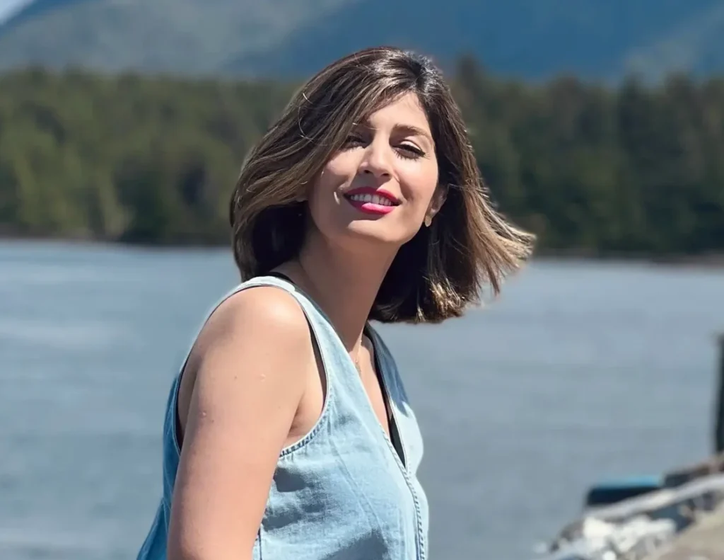 A woman in a light blue denim dress sits on the edge of a weathered wooden dock, smiling toward the camera. She is wearing a sleek, dark grey prosthetic leg and white sneakers. The background shows a large body of water with misty, blue-toned mountains in the distance and a few blurred figures walking further down the dock.