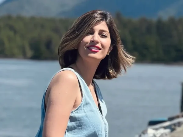 A woman in a light blue denim dress sits on the edge of a weathered wooden dock, smiling toward the camera. She is wearing a sleek, dark grey prosthetic leg and white sneakers. The background shows a large body of water with misty, blue-toned mountains in the distance and a few blurred figures walking further down the dock.