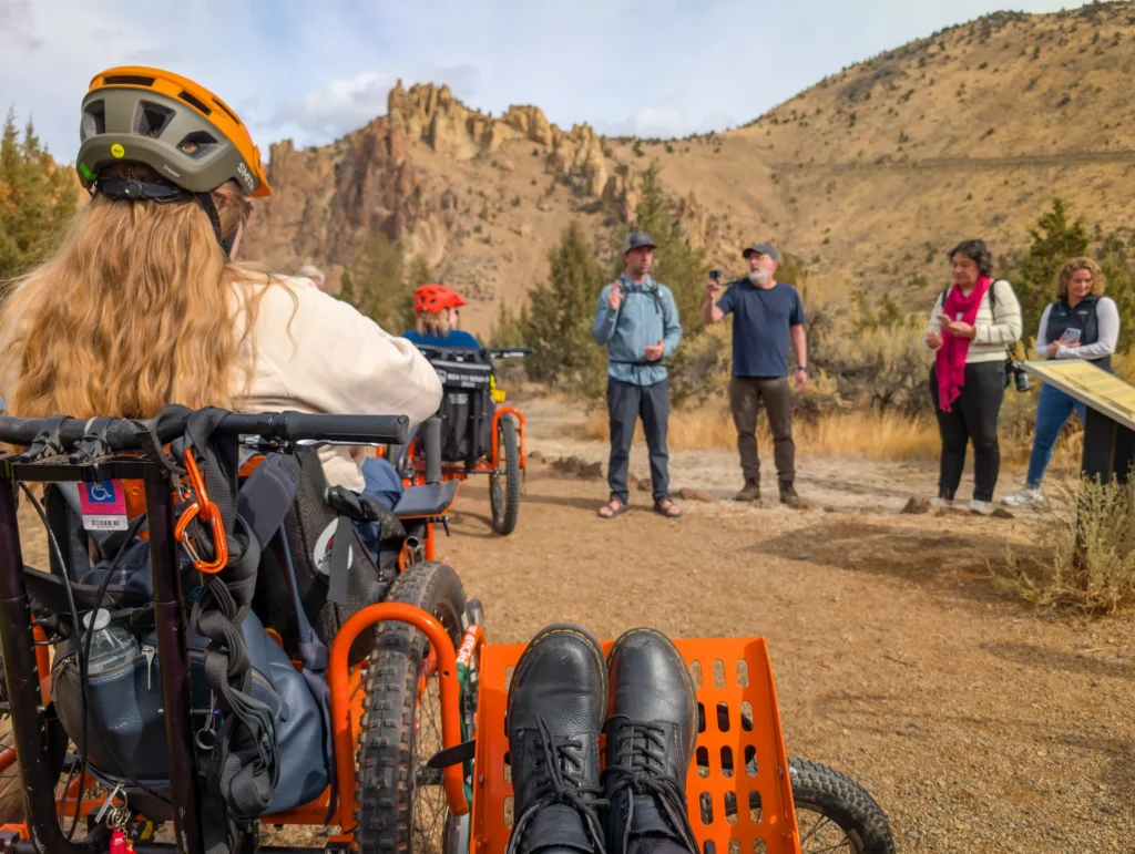 A first-person view from a person sitting in an orange adaptive cycle, showing their black boots resting on the footplate. In front of them, a group of people and another cyclist are gathered on a dirt trail for a briefing, with desert hills and a rocky outcropping in the distance.