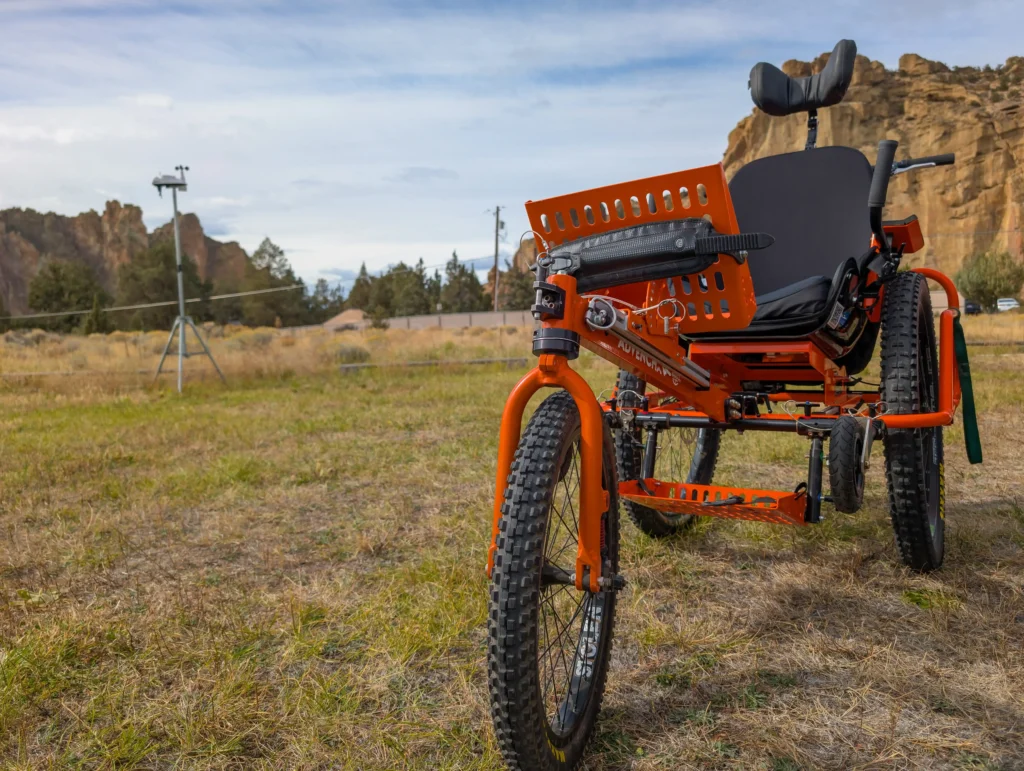A close-up of a specialized bright orange three-wheeled adaptive mountain bike parked on a grassy field. It features heavy-duty off-road tires and a black bucket seat, with a rocky desert cliff and a clear sky in the background.