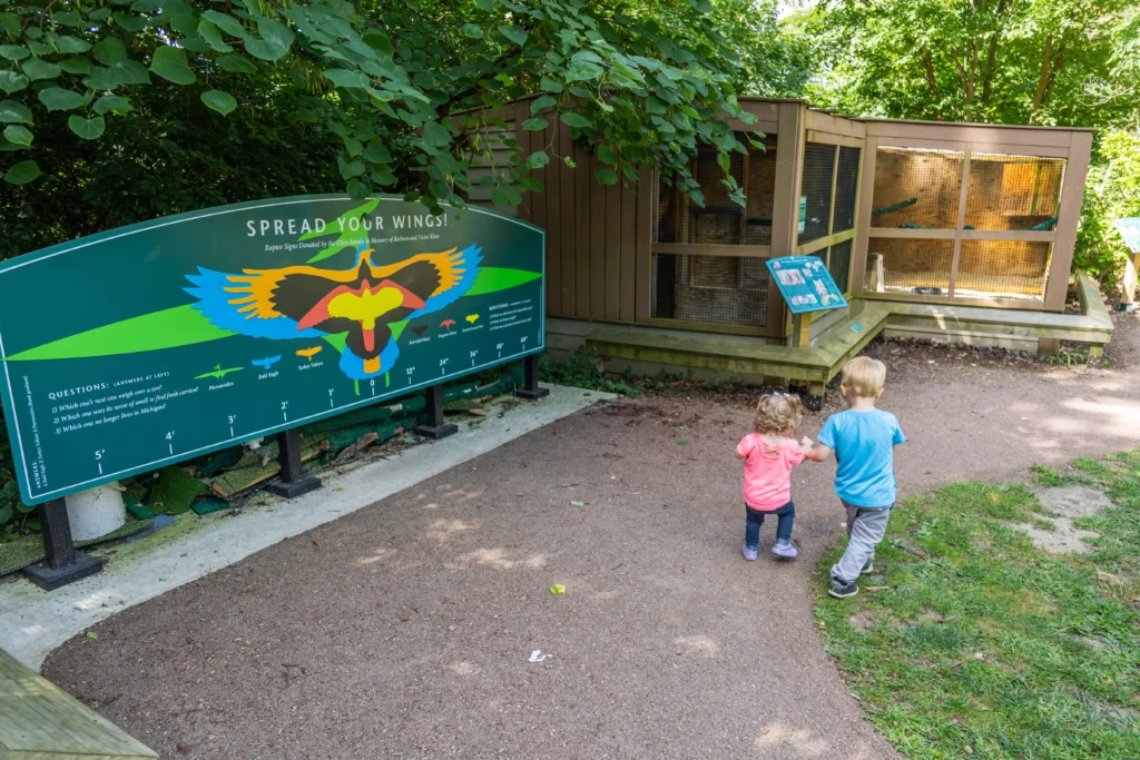 Two young toddlers, a girl in pink and a boy in blue, walk hand-in-hand down a gravel path toward a large green educational sign titled "Spread Your Wings!" The sign shows a colorful diagram of bird wingspans. In the background, there is a wooden raptor enclosure nestled among lush green trees.