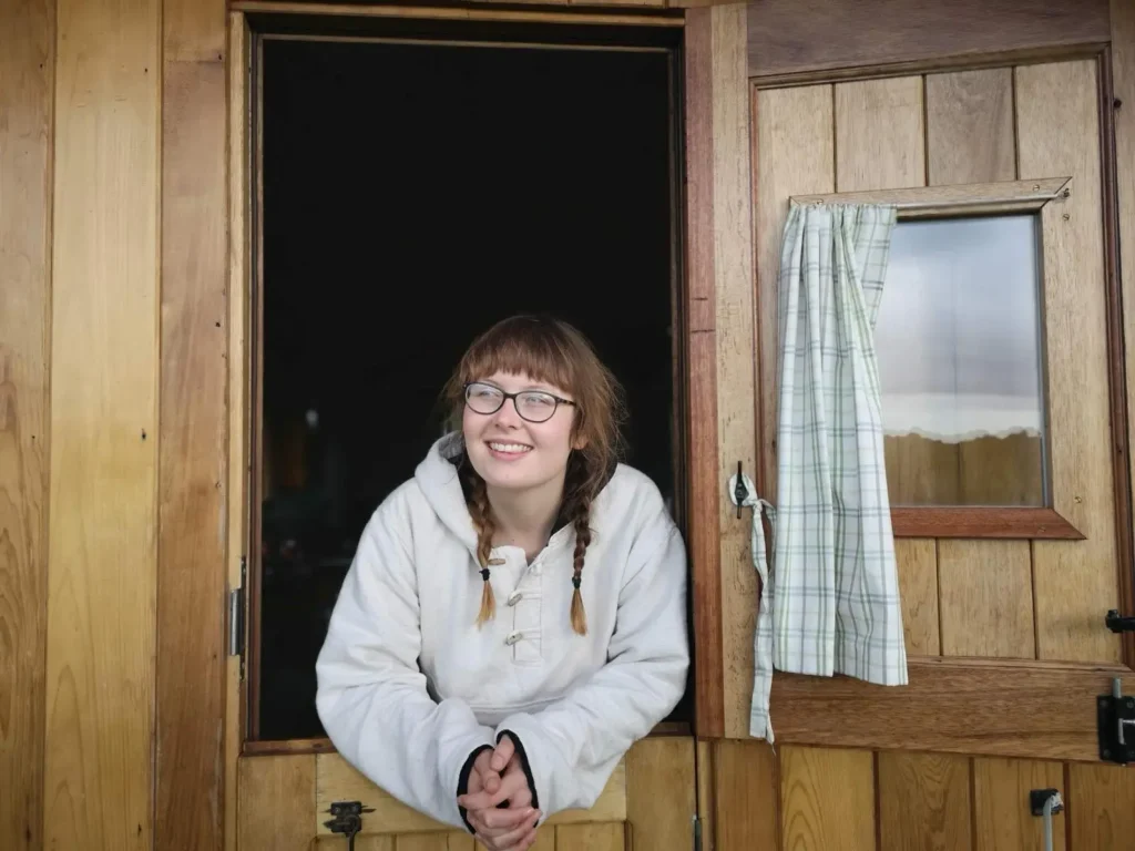 A young woman with red hair in pigtail braids and glasses smiles warmly while leaning out of the top half of a rustic wooden stable-style door. She is wearing a cream-colored hooded sweatshirt.