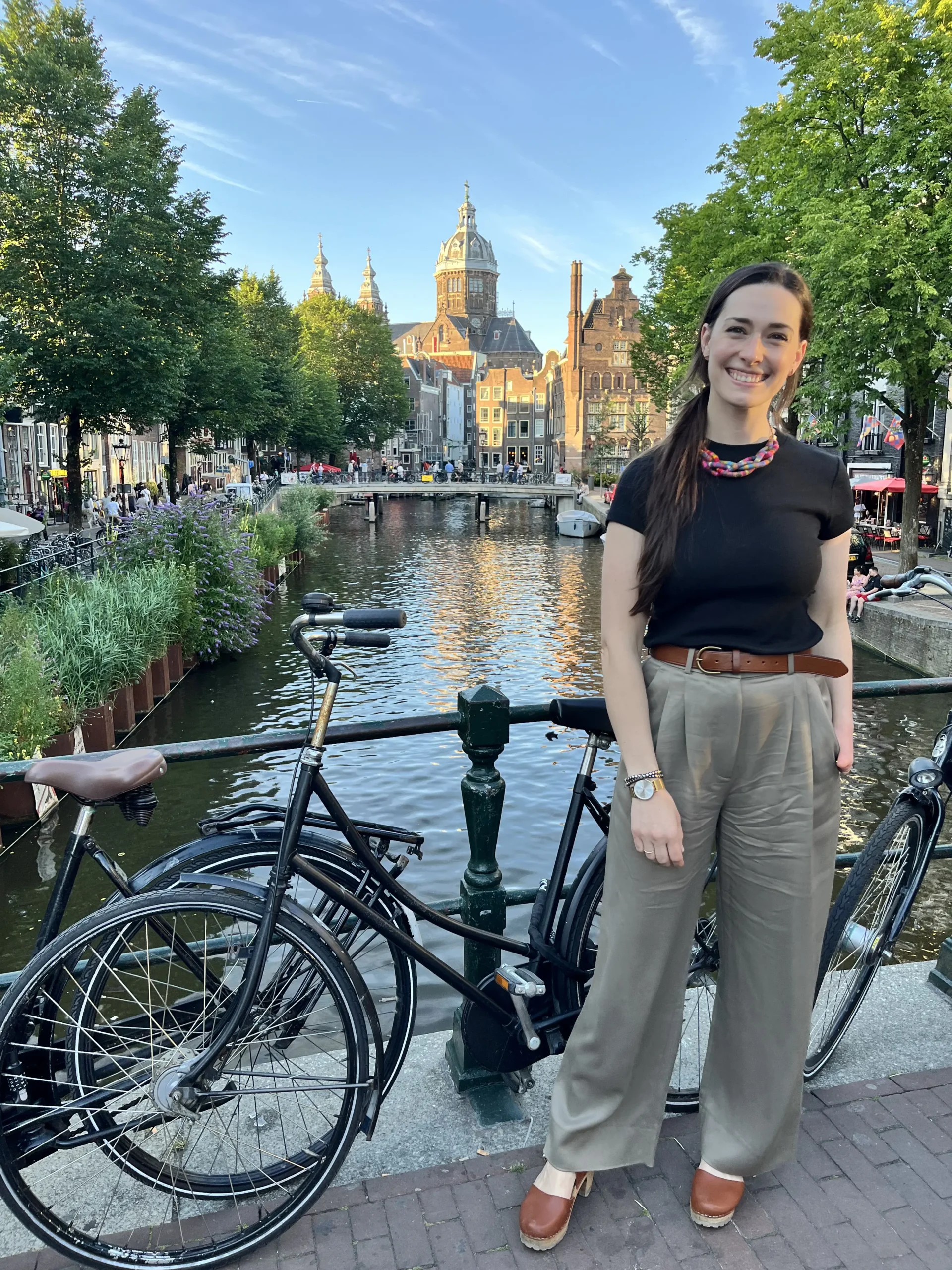 A full-length photo of a woman standing on a brick bridge in Amsterdam. She is wearing a black t-shirt, olive-colored trousers, and a colorful necklace. Behind her are two bicycles parked on the bridge, a scenic canal, and historic Dutch architecture leading toward a large domed church.