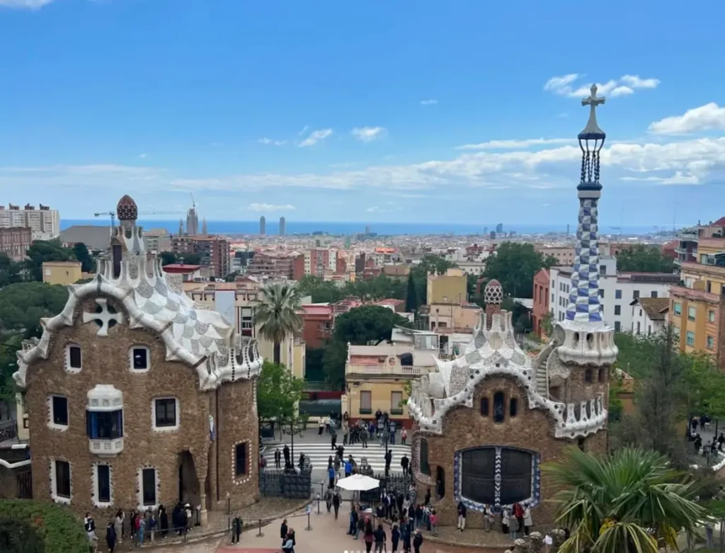 A high-angle scenic view of Park Güell in Barcelona. In the foreground are two distinct gingerbread-style gatehouses with mosaic-tiled roofs and ornate spires. In the background, the dense cityscape of Barcelona stretches toward the blue horizon of the Mediterranean Sea under a clear sky with light clouds.
