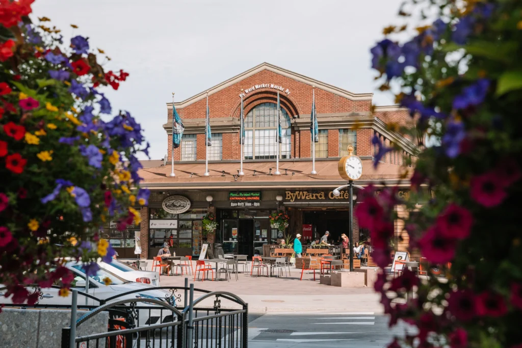 A vibrant daytime view of the historic brick ByWard Market building in Ottawa, framed by colorful purple and red hanging flower baskets in the foreground. The scene features an outdoor patio with red and grey chairs where people are sitting. A classic pedestal clock stands near the entrance under a clear, pale sky.