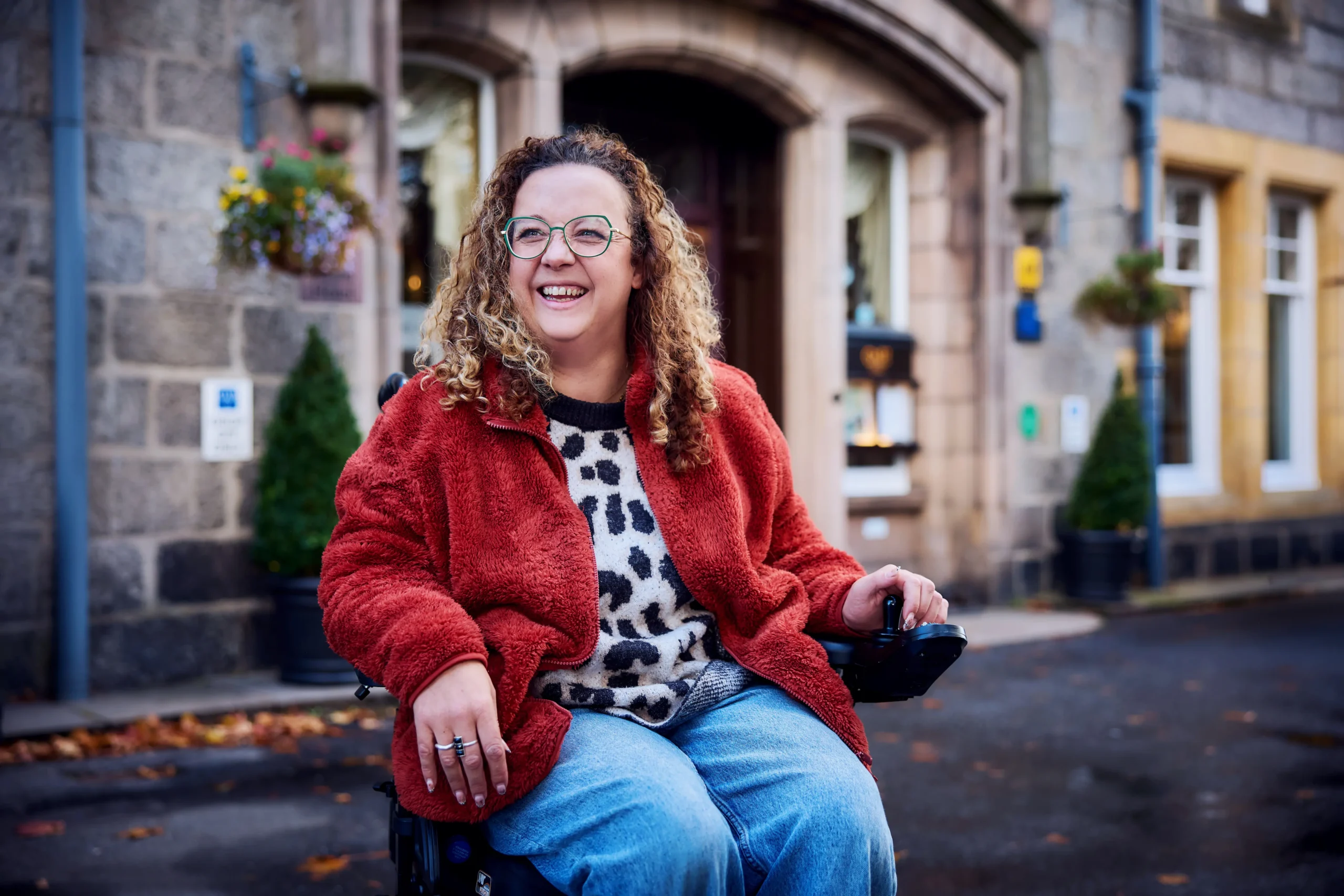 A candid, vibrant photo of a woman laughing while sitting in a power wheelchair outside a stone building. She is wearing a red fleece jacket over a leopard-print sweater and blue jeans. Small orange leaves are scattered on the ground around her.